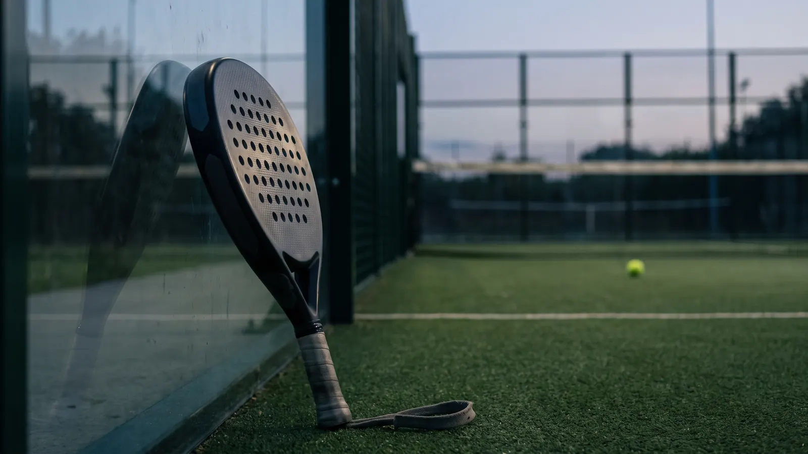 close-up of a diamond-shaped solid perforated carbon padel racket resting against the glass wall of a court, textured face, soft evening lig