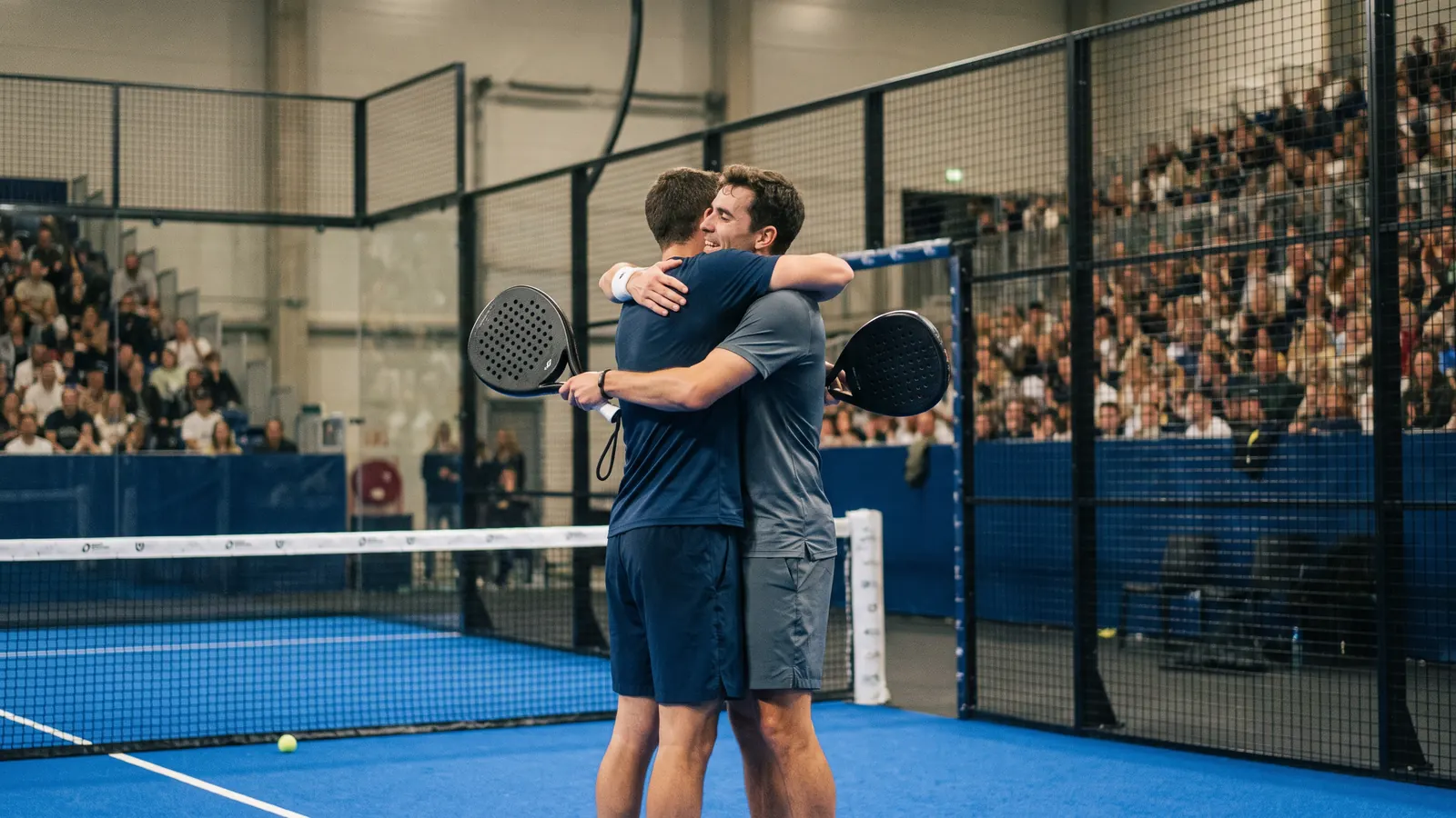 two padel players embracing in celebration on a blue glass-walled indoor padel court in a packed arena under bright lights, solid perforated