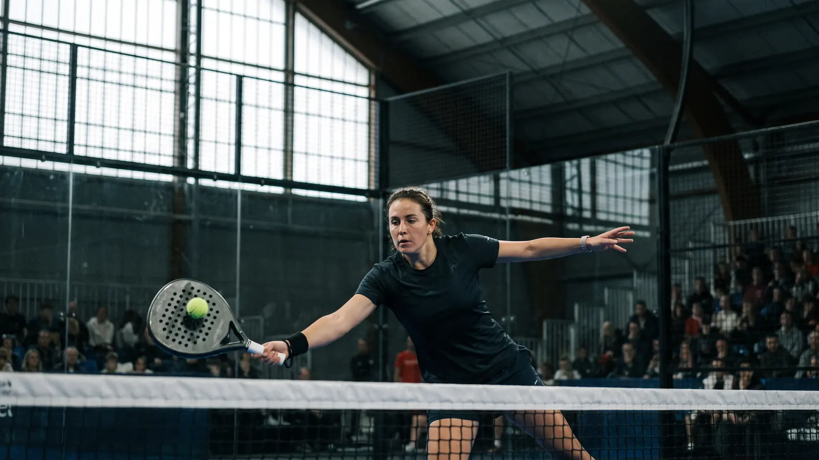 a female padel player hitting a forehand volley at the net on an indoor glass padel court in a large arena, crowd blurred in the background,
