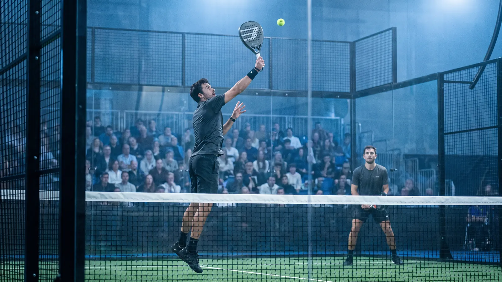a padel doubles match in a packed indoor arena under bright blue lighting, a player hitting a high overhead near the net, panoramic glass wa