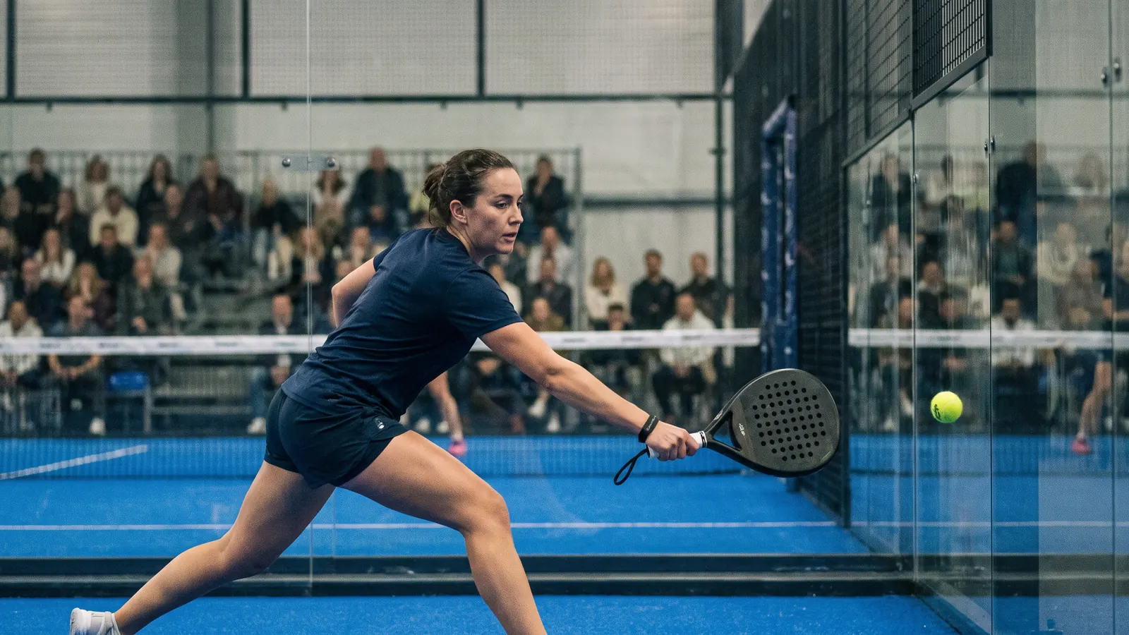 a padel player stretching to return a ball near the back glass in a large indoor arena under bright lights, crowd softly blurred in the stan