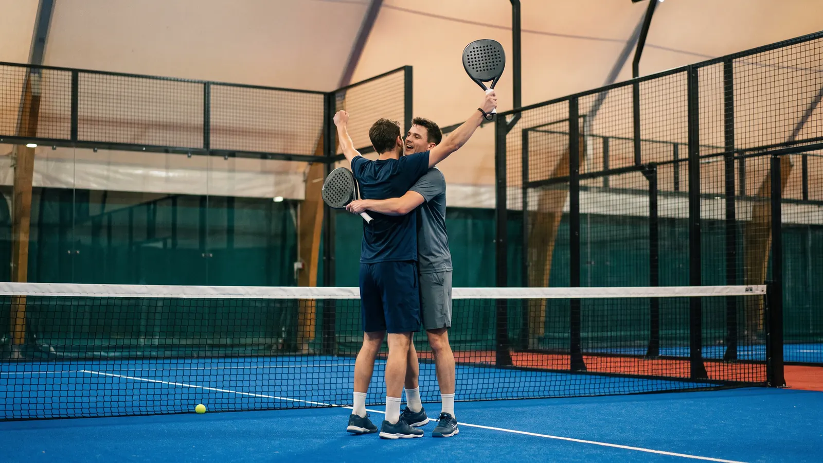 two padel players celebrating a hard-won point at the net on a blue glass court in a big indoor arena under bright lights